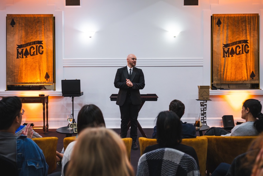 A seated audience in an intimate theatre room setting watching a magician perform.