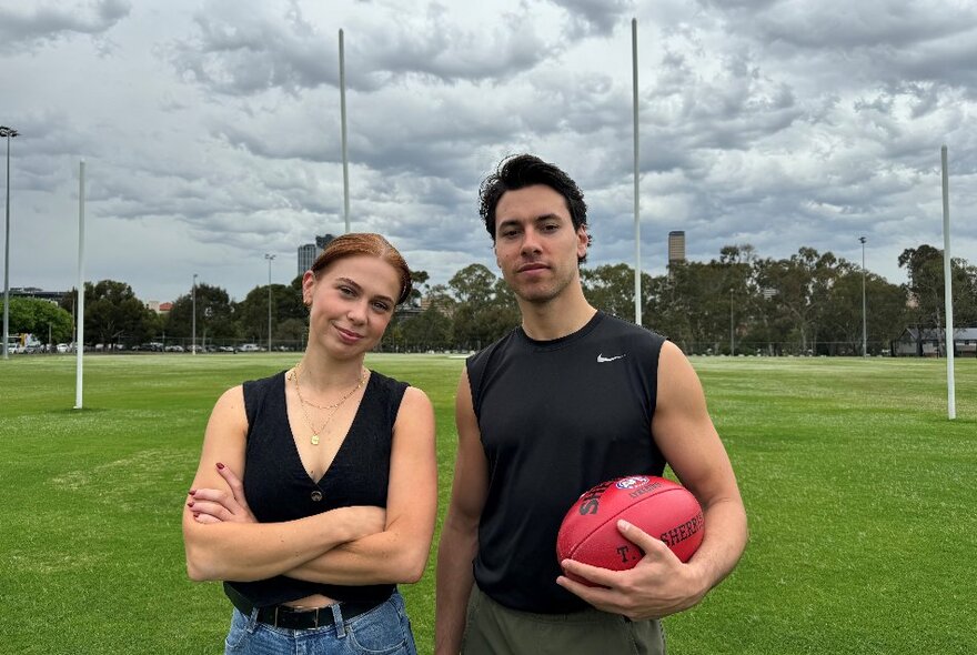 Two actors standing on a football pitch, the male holding a red football, female with arms crossed, goal posts in the background.