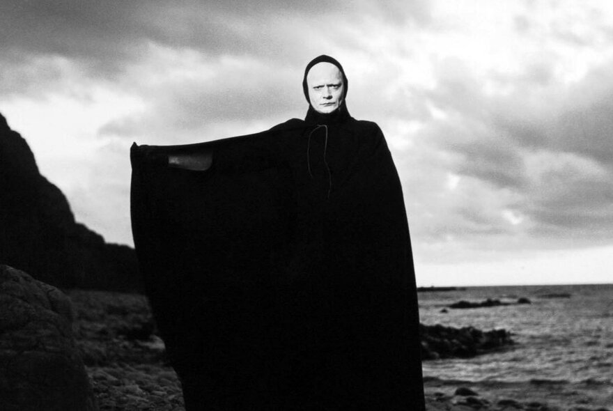A still image from the film The Seventh Seal, showing a man in white face paint in a black robe by the sea, one arm stretching the robe out sideways. 