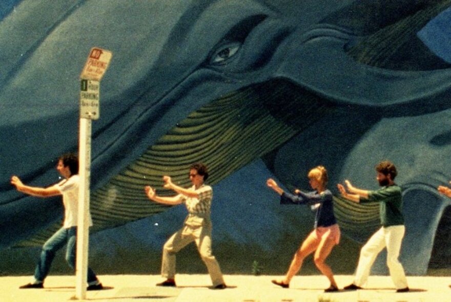 A still from the film, Mur Murs, depicting four people practicing T'ai Chi in front of a giant mural painting of a blue whale.