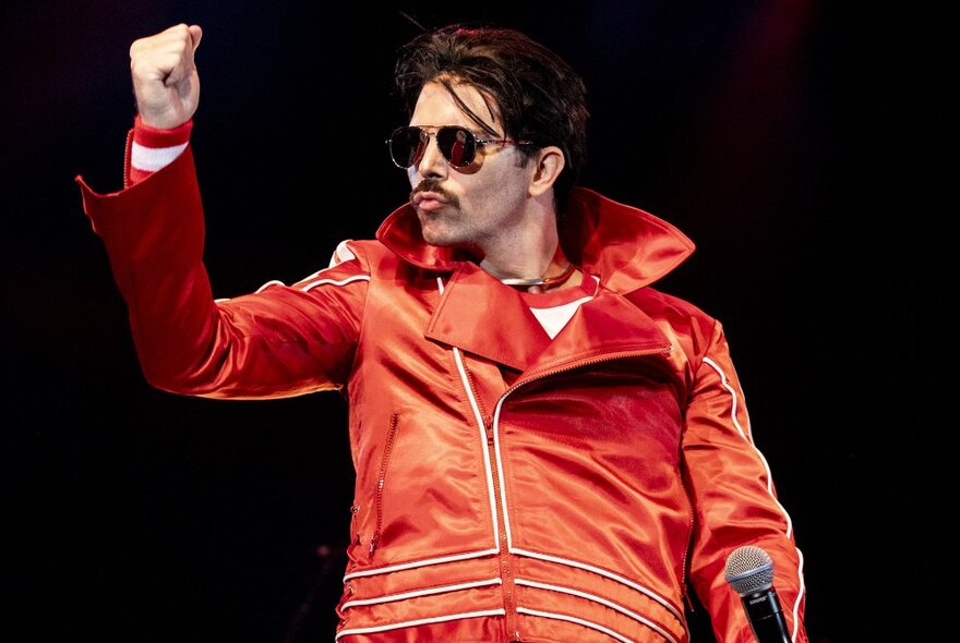 A tribute performer on stage dressed as Freddie Mercury in a red satin jumpsuit with large collar.