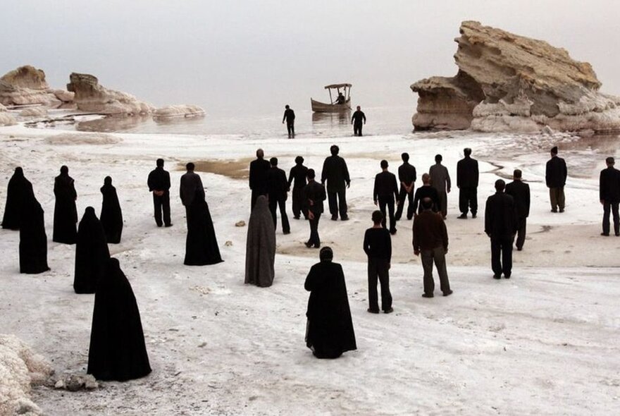 A still from a film by Mohammad Rasoulof showing black-robed figures on a pale, ghostly beach with a small boat in the water.