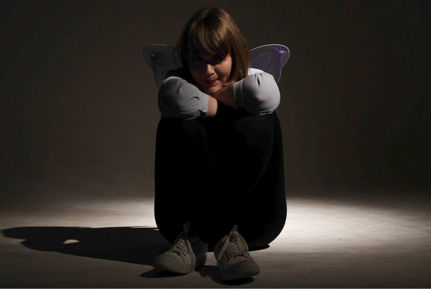 An actor seated on the ground, her legs tucked up and her arms resting on her knees, sitting in semi-darkness.