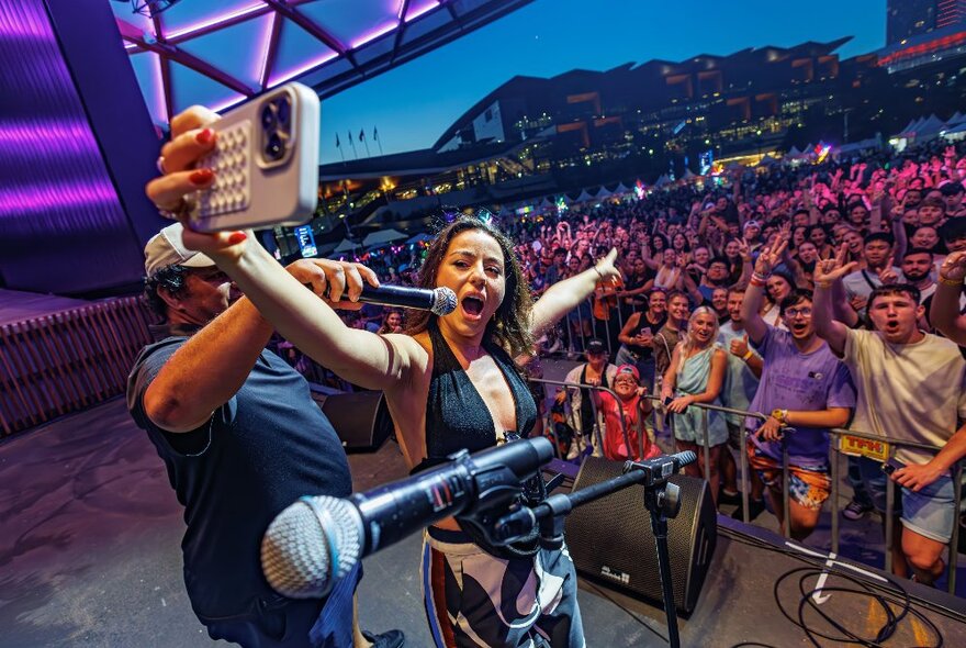 A performer on stage takes a selfie with a large crowd behind her, microphones in the foreground. 