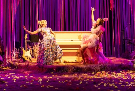 Two actors in costume performing on a small stage in front of a piano and velvet curtain.