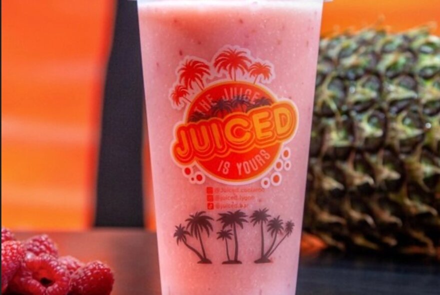 A takeaway plastic cup of fresh pink coloured juice, with branding on the cup, and raspberries and a pineapple next to it.