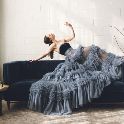 A performer dressed in a dramatic ruffled grey gown leaning back on the top edge of a couch in a stylised pose, one arm raised in the air.