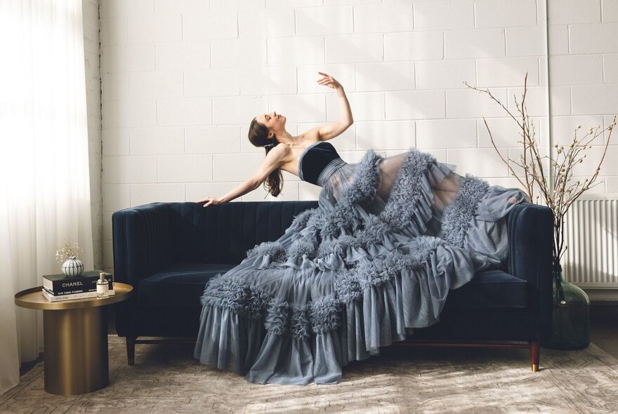 A performer dressed in a dramatic ruffled grey gown leaning back on the top edge of a couch in a stylised pose, one arm raised in the air.