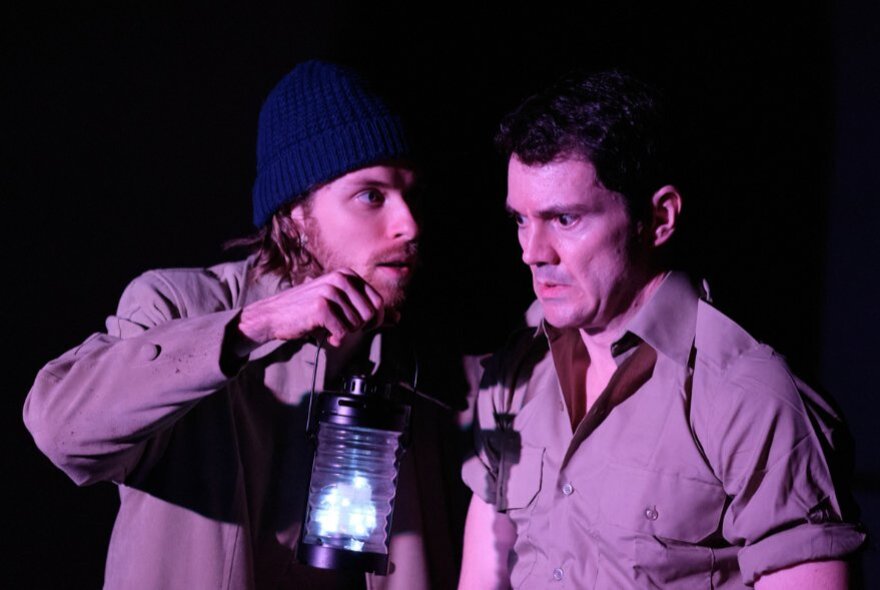 Two actors on a stage, one holding a lantern, both looking serious.