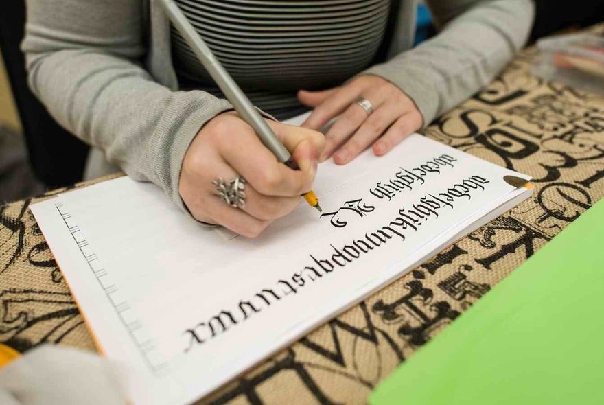 A person's hand holding an ink pen and writing calligraphy on a large white journal.