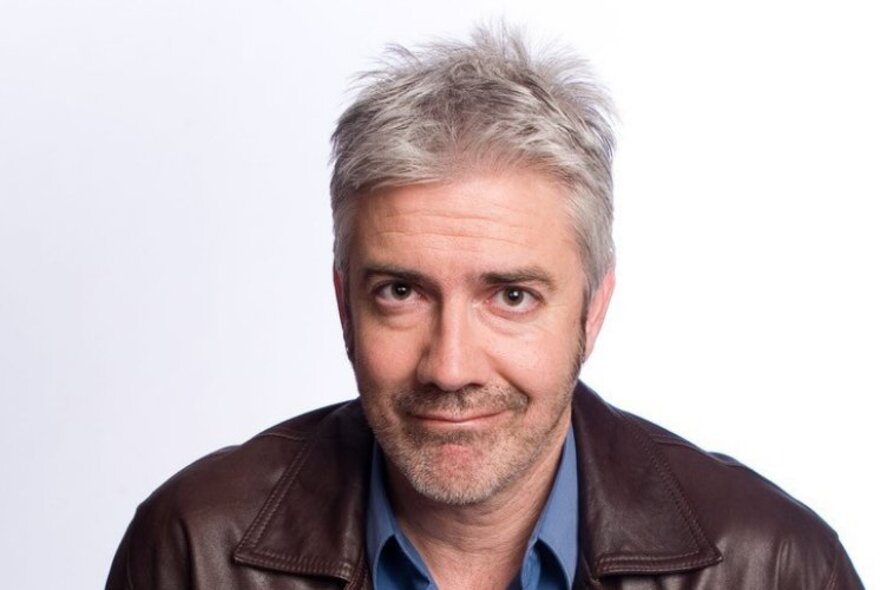 Portait of renowned Australian comedian, Shaun Micallef, wearing a leather jacket and blue shirt, smiling.