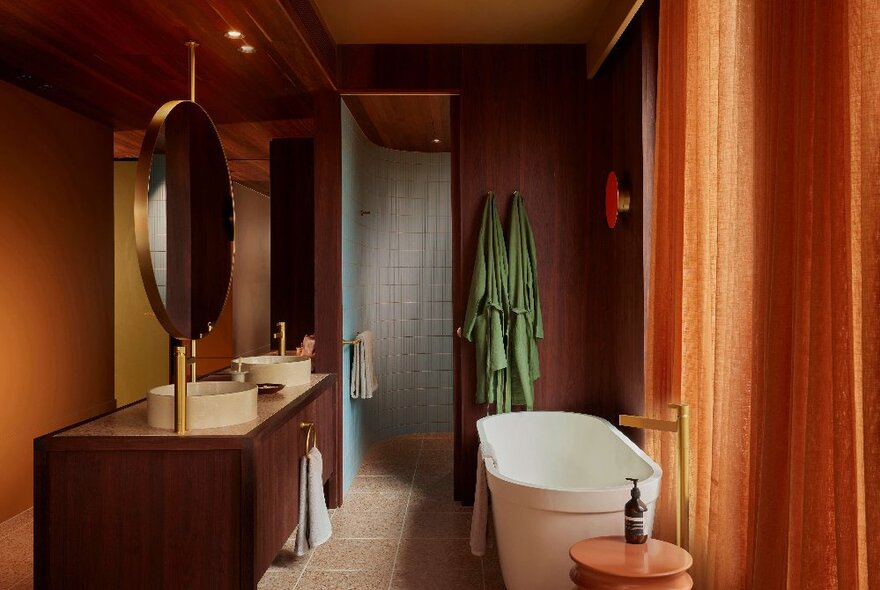 An orange toned hotel bathroom with a white tub and round mirror, a green bathrobe hangs at the back. 