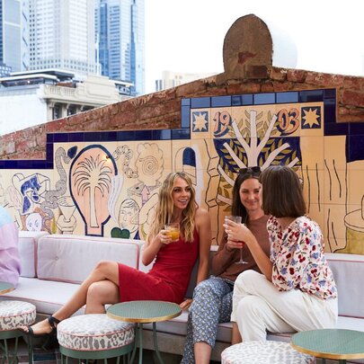 People seated on banquette couches at an open air rooftop bar with the Melbourne skyline in the background.