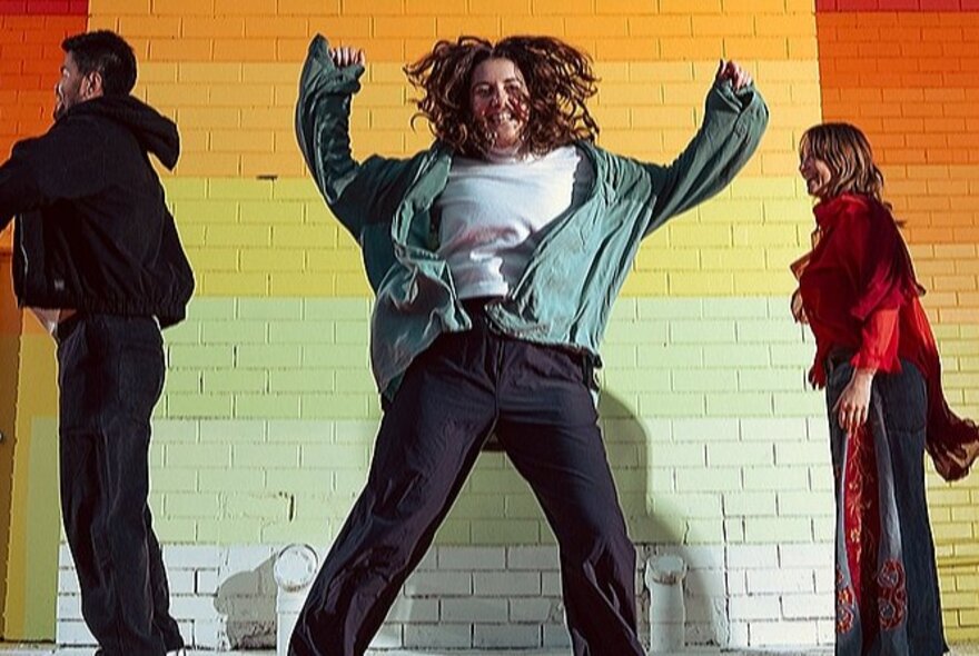 Three people dancing with a rainbow painted brick wall behind them.