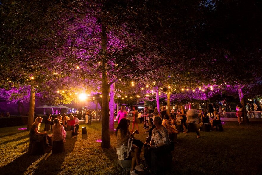 The garden at NGV International, festooned with hanging lights under trees, with people seated on stools on the lawns enjoying music and food.