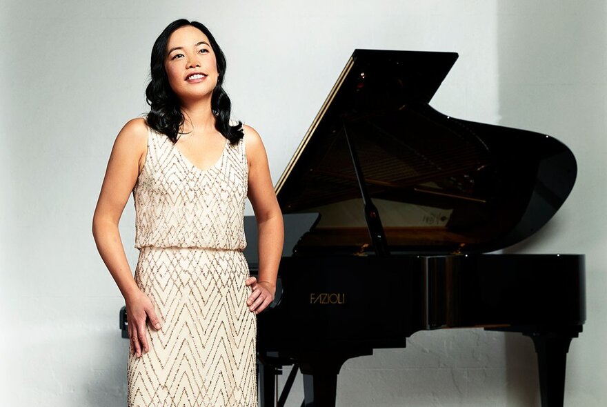 Pianist Andrea Lam wearing a sleeveless shimmering pale dress standing in front of a grand piano.