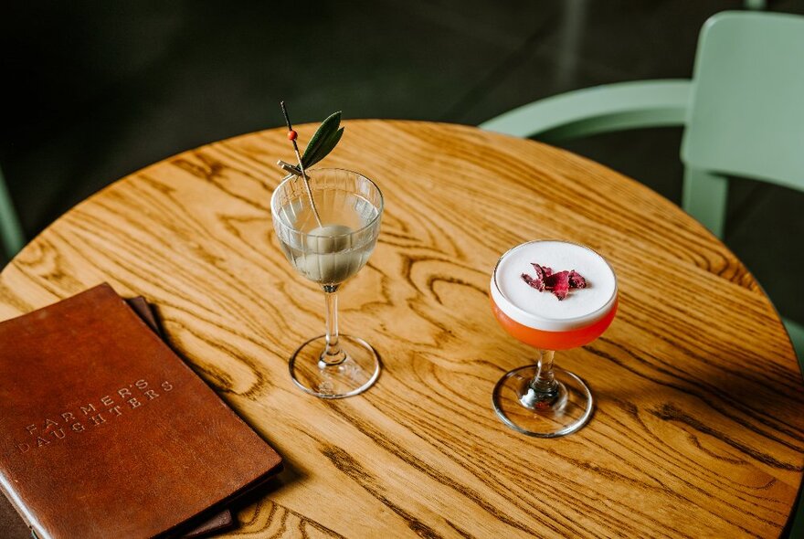 Two cocktails and a leather bound menu on a round wooden table in a restaurant.