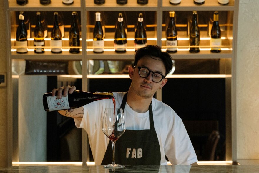 A waiter wearing an apron that reads FAB pours a single glass of red wine at a bar. 