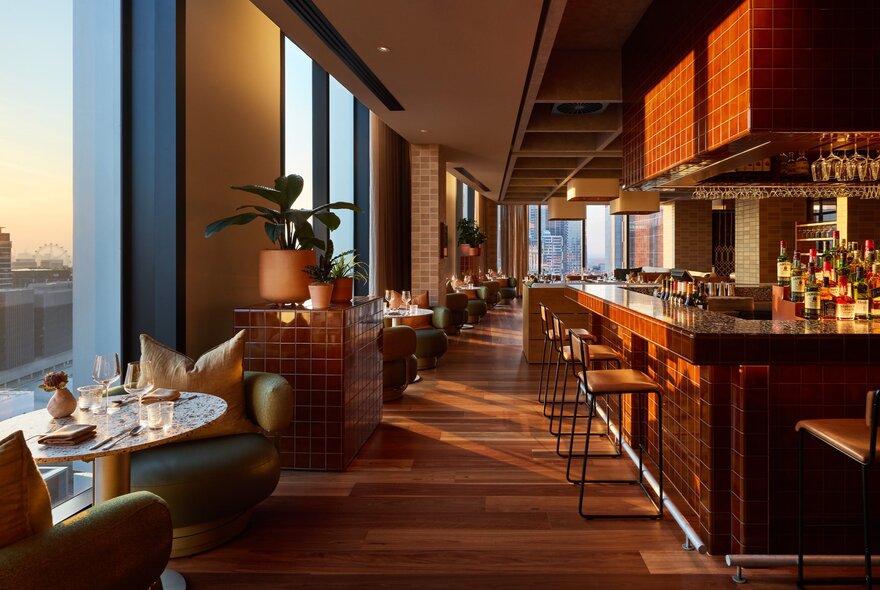 A modern rooftop restaurant interior with a long wooden bar, barstools, warm lighting, and dining tables next to large windows overlooking a city skyline.