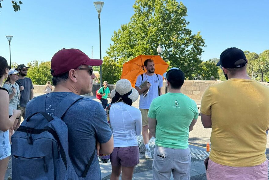 A guide holding an orange umbrella standing on a city street and talking to a small group of people on a walking tour of the city.