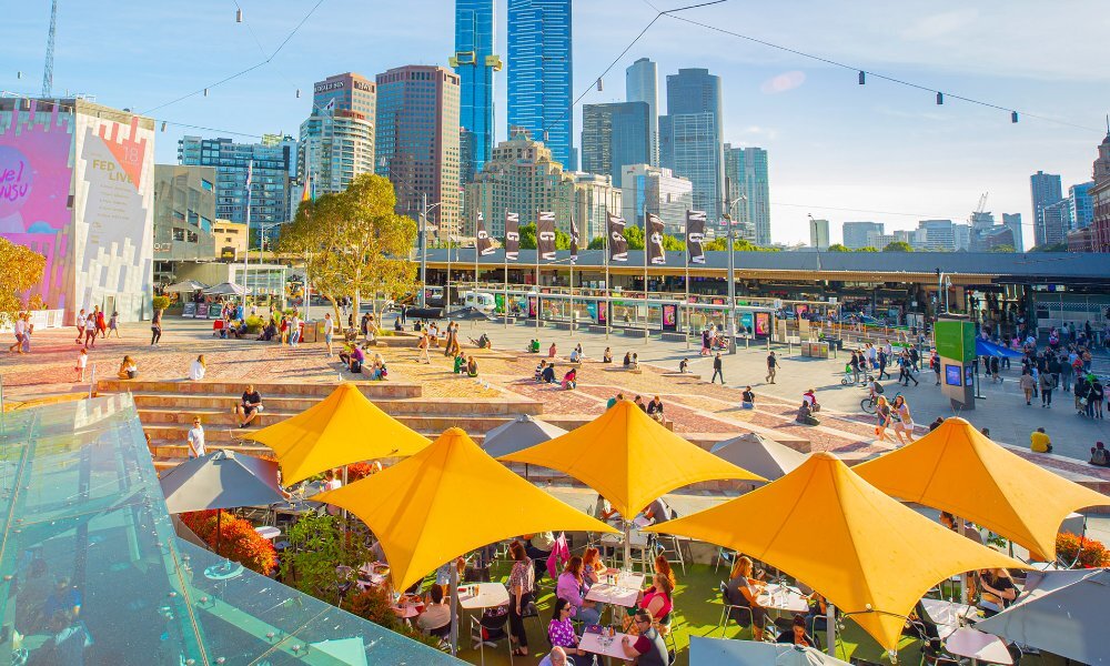 Crowds dining under yellow umbrellas in Fed Square on a sunny day.