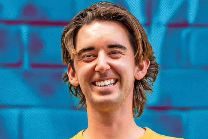 A man with shaggy hair laughing against a blue brick background. 