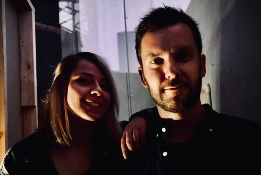 The head and shoulders of a man and a woman standing close together in a dimly lit room slightly smiling at the viewer.