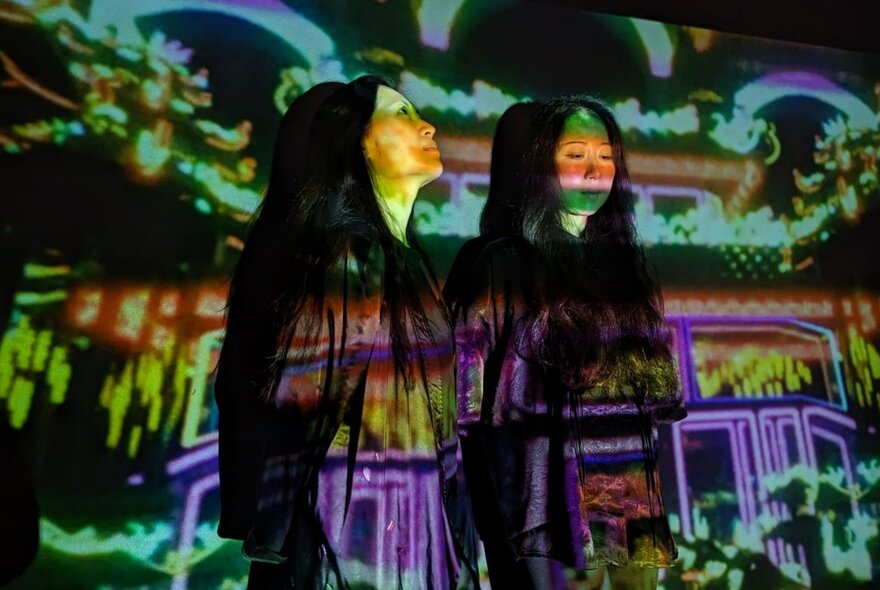 Two women with long dark hair standing together in a room with colourful projections on them and their surrounds.