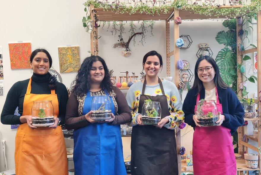 Four smiling participants in a studio workshop setting, wearing colourful aprons and holding glass-domed terrariums.