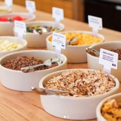 An array of toppings for frozen yoghurt in white bowls displayed on a wooden counter.