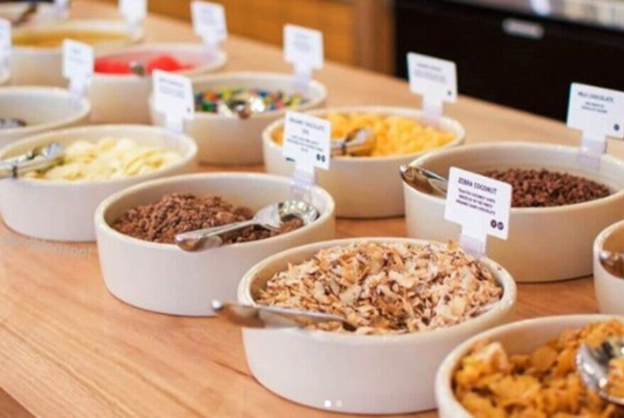 An array of toppings for frozen yoghurt in white bowls displayed on a wooden counter.