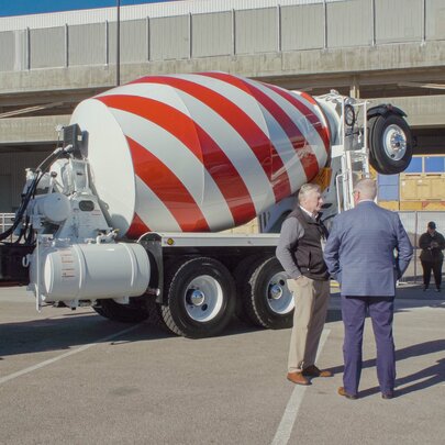 A still from the documentary film, The History of Concrete, of a cement mixer truck with a red and white striped barrel parked on a paved lot, with two men standing in conversation nearby.