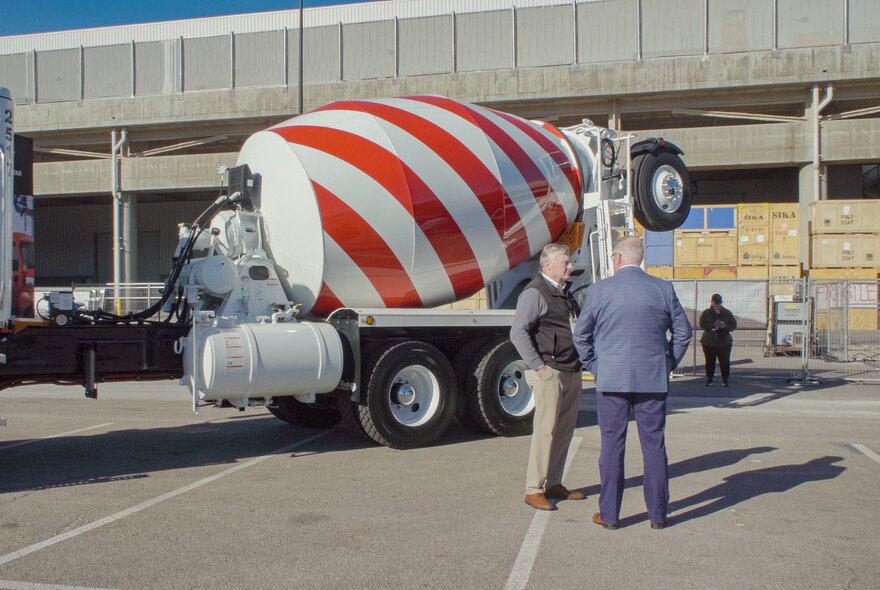 A still from the documentary film, The History of Concrete, of a cement mixer truck with a red and white striped barrel parked on a paved lot, with two men standing in conversation nearby.