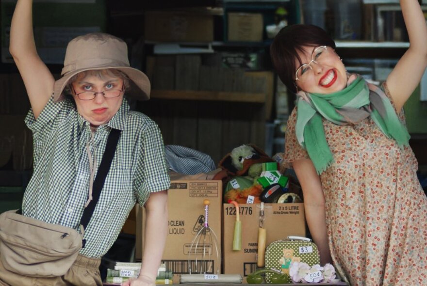 Two performers, one dressed as a male, standing with arms raised beside boxes of items at a garage sale.