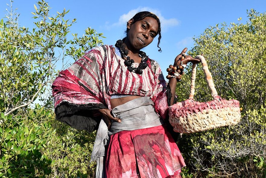 An Indigenous model in a garden holding up a basket with one finger and the other hand on her hip. 