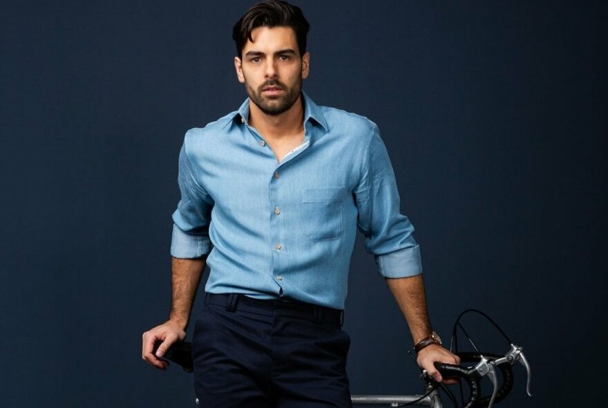 Man in light blue shirt, leaning against racing bike, against dark blue, mottled background.