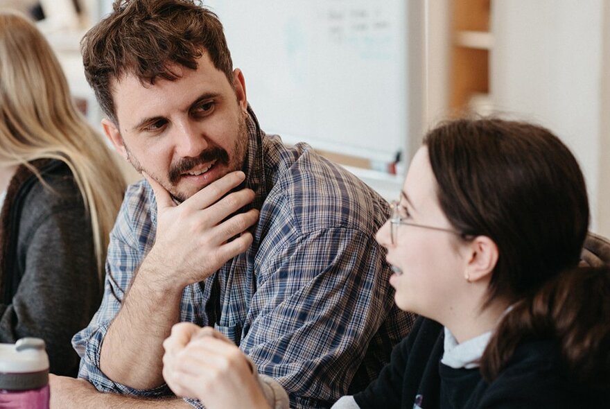 A man with his hand on his chin talking to a young woman in a classroom setting. 