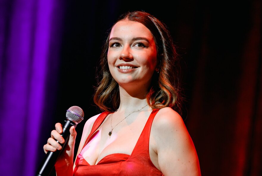 A singer in a red dress, smiling and holding a microphone while on stage, a purple stage curtain behind her.
