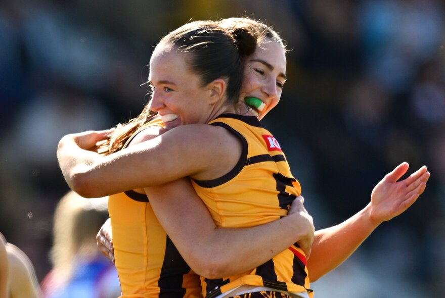 Hawthorn AFLW players hugging and celebrating a moment on the field in their brown and gold guernseys.