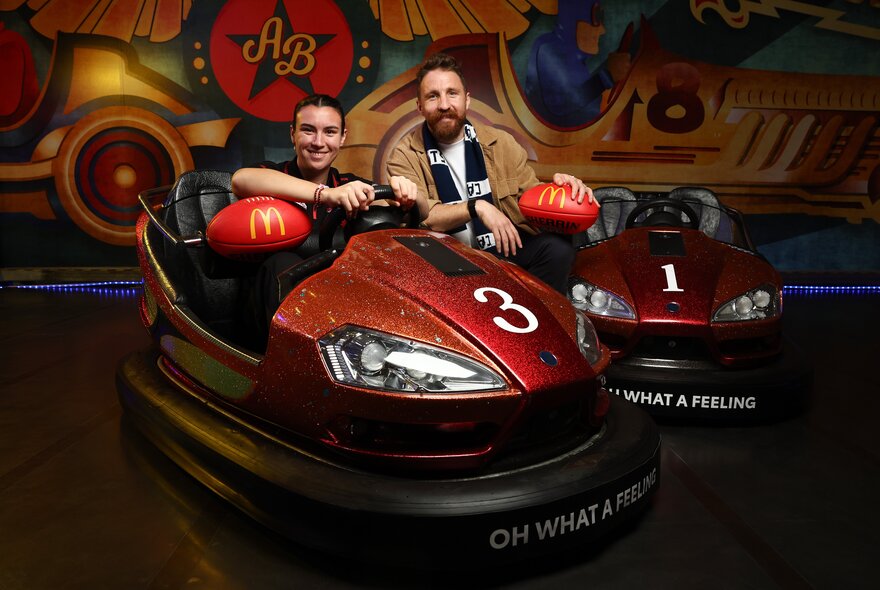 Two AFL celebrities seated inside dodgem cars, both holding red footballs.