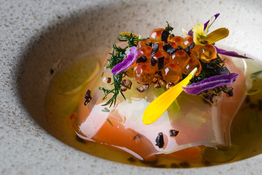 A ceramic shallow bowl filled with clear broth and small portions of food, garnished with flower petals and caviar.