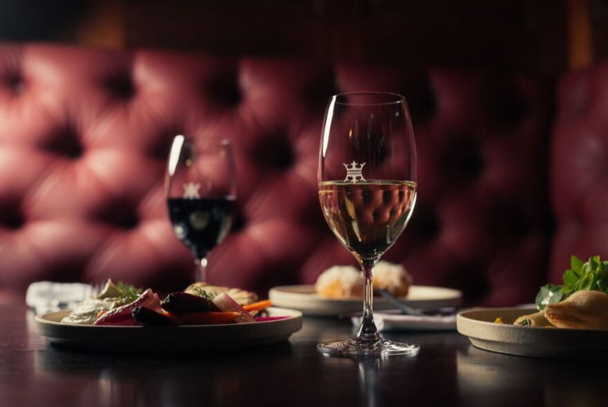 Two plates of food and two glasses of wine served in a restaurant setting, burgundy leather banquettes in the background.