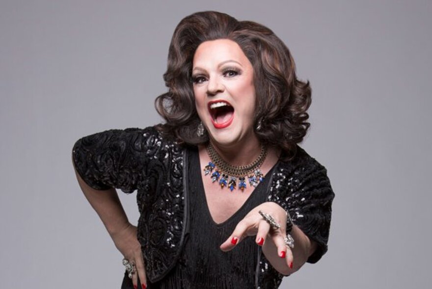 Australian cabaret drag performer Dolly Diamond, with an oversized hairdo, and one  arm reaching out towards the viewer, against a neutral backdrop.