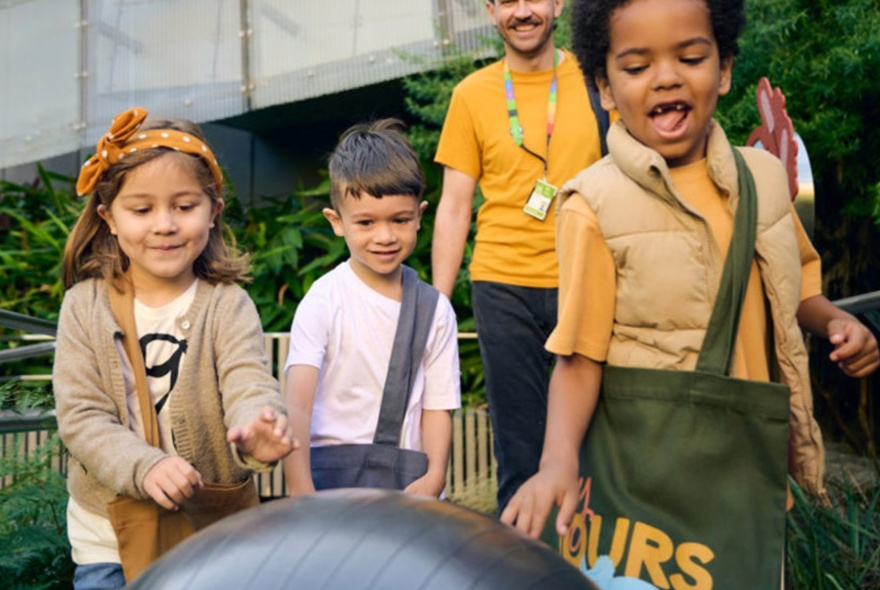 Three children and an adult in the background, engaged in an activity in a garden setting at Melbourne Museum.