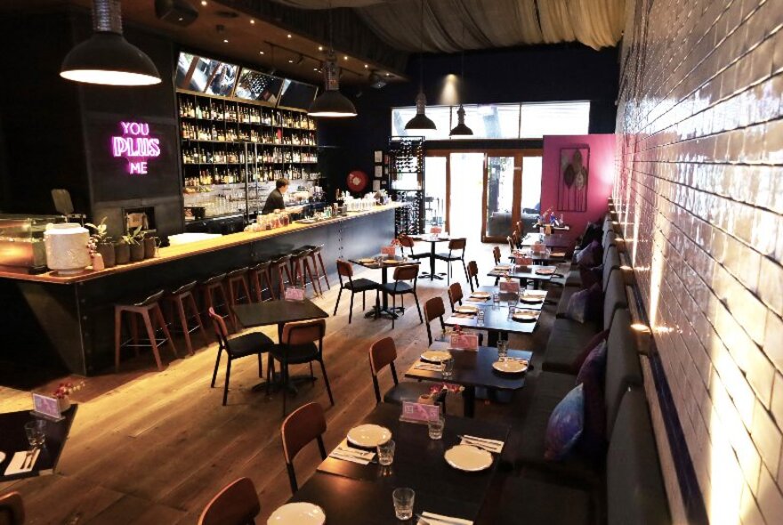 The interior of Plus 5 Bar, with a long bar area on the left and a row of small tables and chairs arranged along a brick wall on the right. 