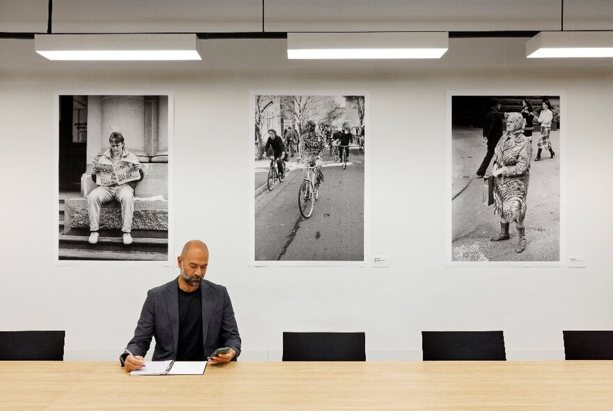 A man working at a table at City Library, writing into a notebook, three large black and white photographs displayed on the wall behind him.