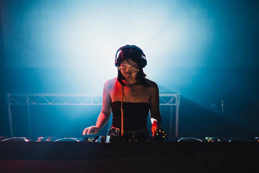 A young woman wearing headphones and playing music on a DJ deck, a blue light behind her.