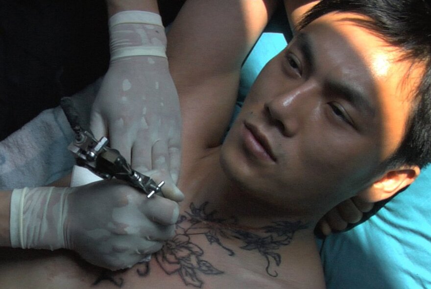 Film still of a man reclining on his back while his chest is being tattooed by a person wearing white surgical gloves holding a tattoo gun.
