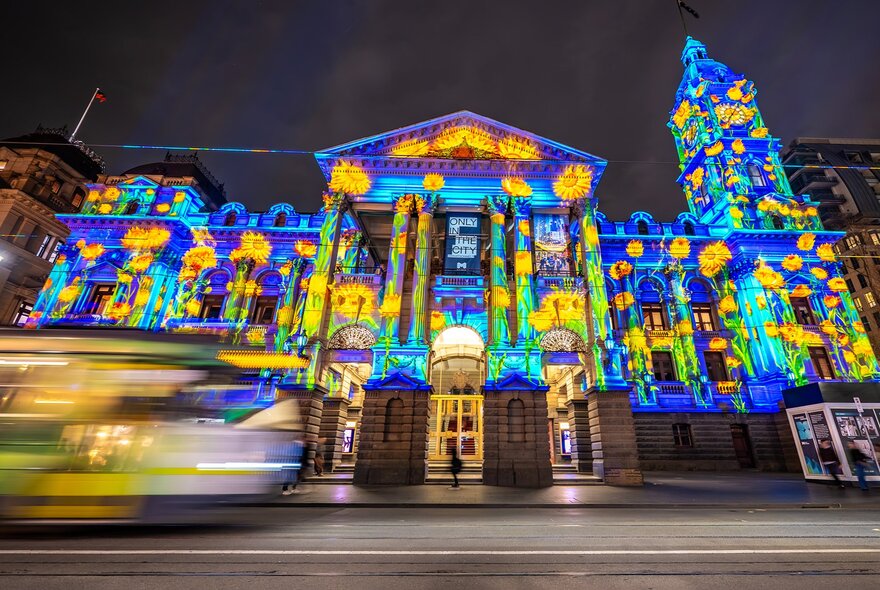 The facade of Melbourne Town Hall illuminated in colours, with a blurred tram going past.