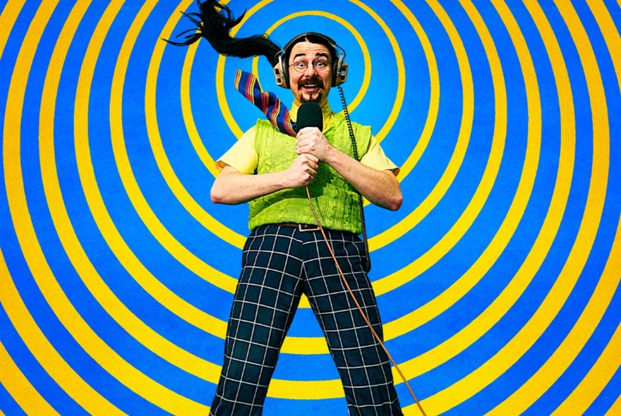 A man with a long ponytail wearing headphones, a tie, a green knitted vest and checked pants, holding a microphone; yellow and blue spiral background.